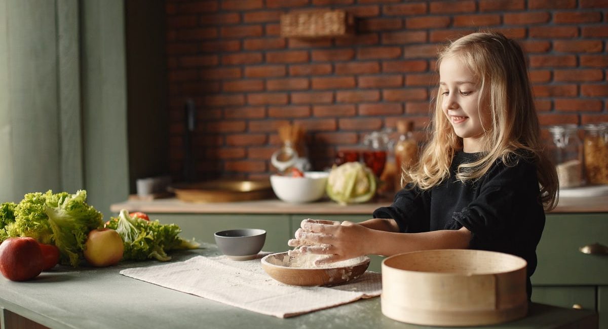 Playful blond child touching flour put into wooden plate while standing near table with fresh vegetables in loft style kitchen in daylight