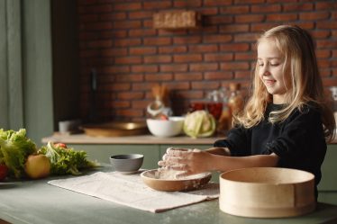 Playful blond child touching flour put into wooden plate while standing near table with fresh vegetables in loft style kitchen in daylight