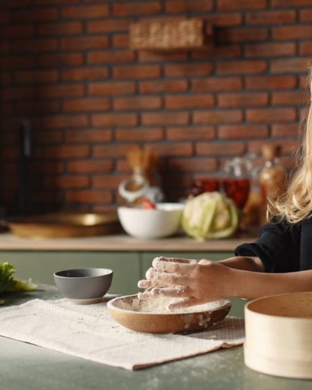 Playful blond child touching flour put into wooden plate while standing near table with fresh vegetables in loft style kitchen in daylight
