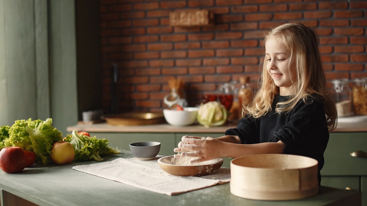 Playful blond child touching flour put into wooden plate while standing near table with fresh vegetables in loft style kitchen in daylight