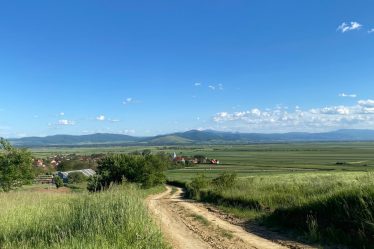 A scenic view of a dirt road leading through fields and hills in Angheluș, Romania under a clear blue sky.
