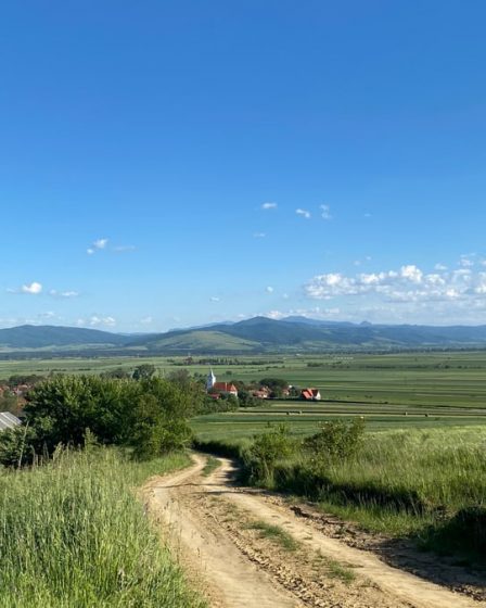A scenic view of a dirt road leading through fields and hills in Angheluș, Romania under a clear blue sky.
